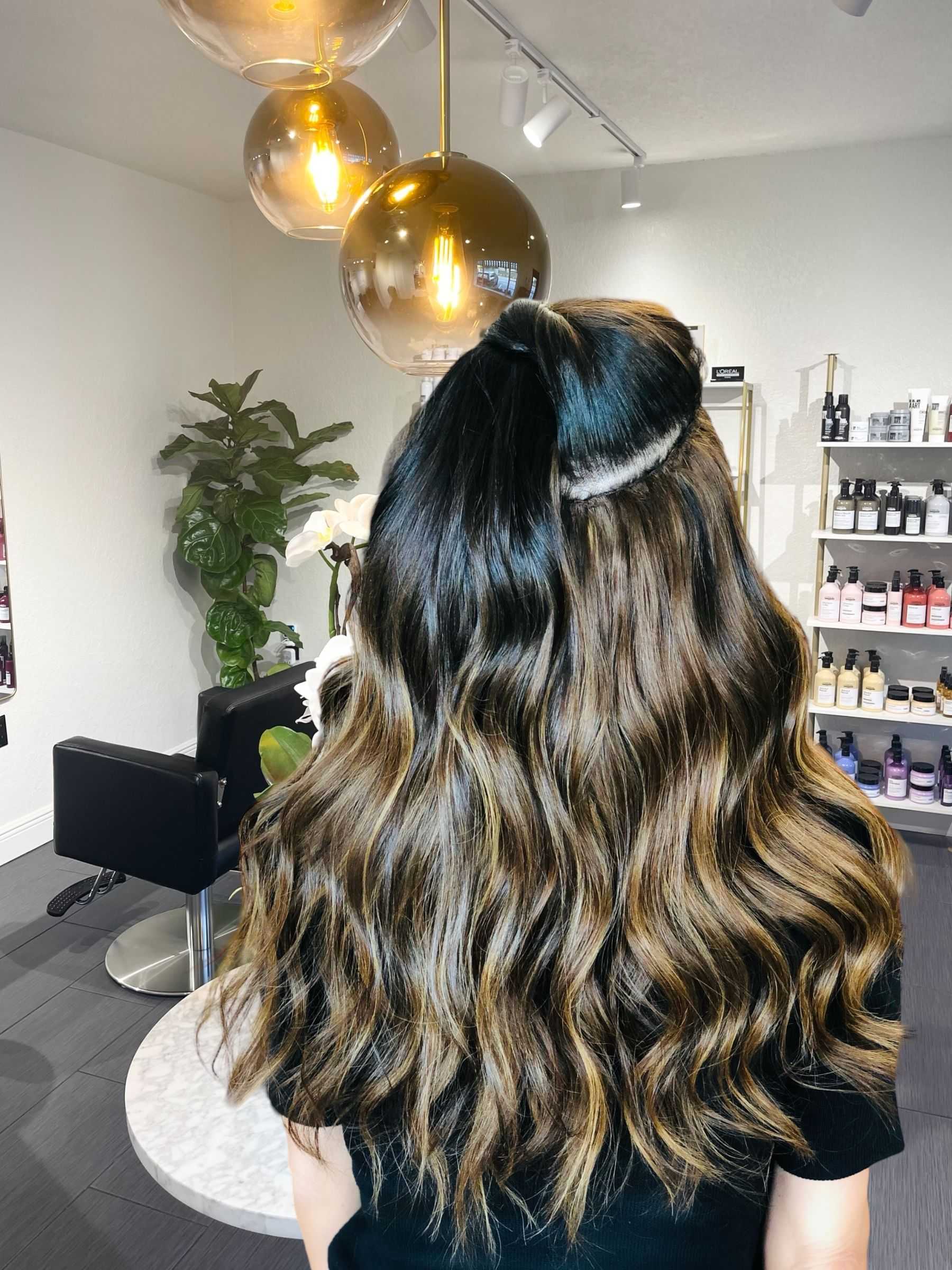 Woman with styled long wavy hair in a modern salon with hanging lights and hair products display.